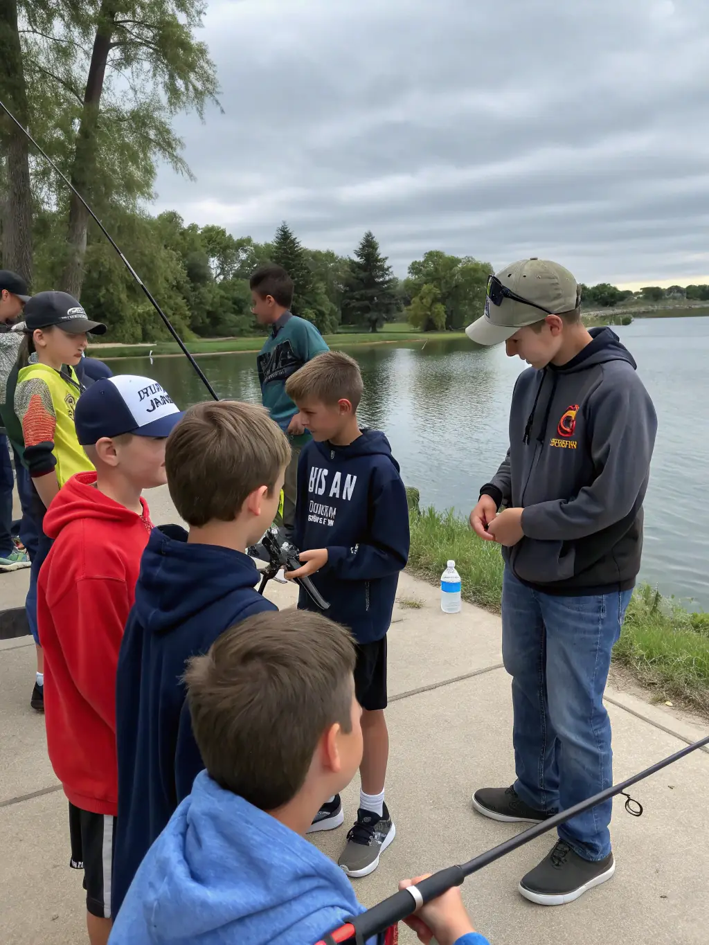 A group of children learning how to cast fishing rods at a TEAM CARNA fishing school, with instructors providing guidance and support. The setting is a calm lake with lush greenery in the background.